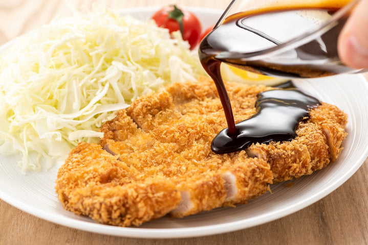 Tonkatsu sauce being poured over a pork cutlet. A cabbage salad with cherry tomatoes is on the side.