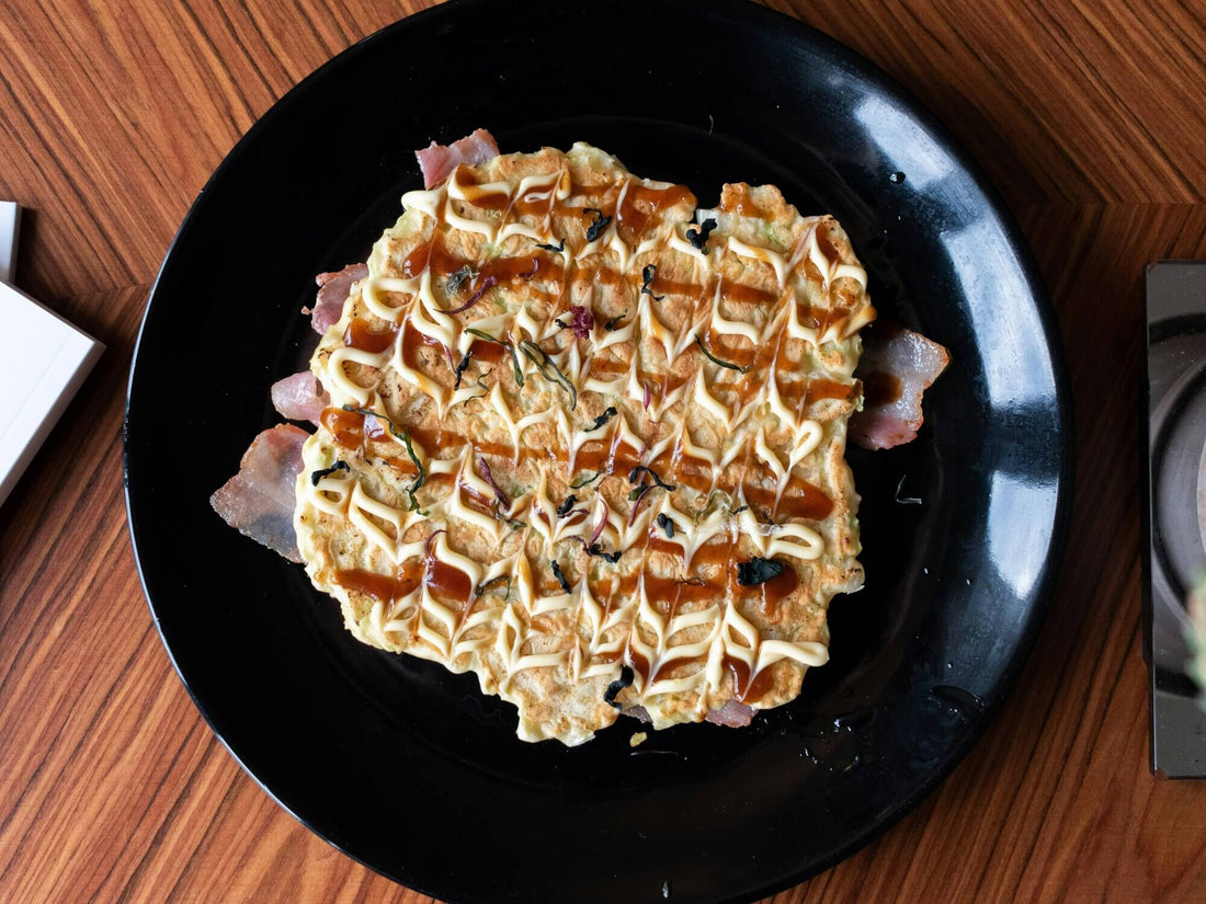 A black plate with Okonomiyaki over bacon sits on a table