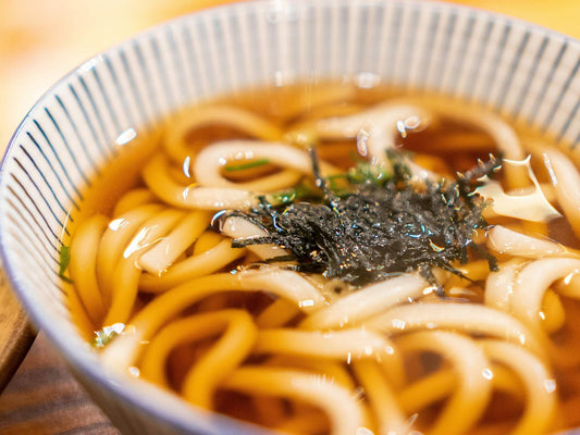 A bowl of udon noodles in a brown soup with seaweed on top